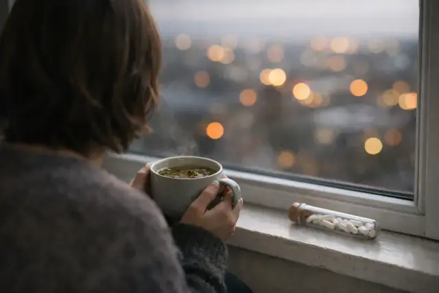 Personne de dos en pull gris tenant une tasse fumante devant une fenêtre, un tube de granules blancs sur le rebord, lumières de ville floues